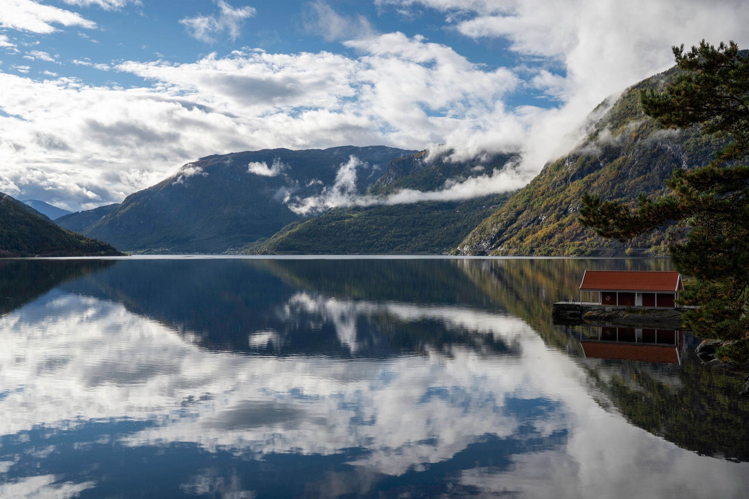 Montañas y nubes reflejadas en un fiordo noruego junto a una pequeña cabaña roja